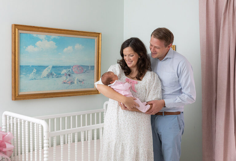 Mother and father in a baby nursery in front of the crib during newborn photography session.  Mom is holding the newborn and both parents are sweetly looking at the child.