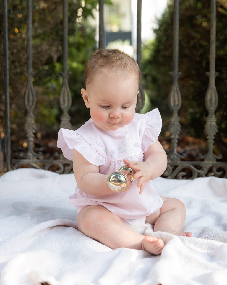 Baby holding a silver rattle while in front of a gate during a professional photography session.  She is wearing a classic pink smocked outfit.