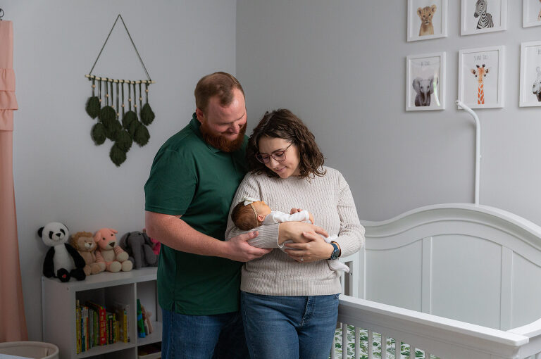 Parents stand in newborn nursery during professional photography session in their home. Standing in front of the crib at holding the newborn lovingly in their arms, Huntsville in Madison County newborn photographer.