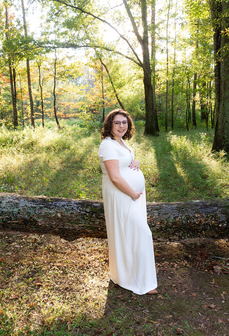 Pregnant woman with a white dress standing in a green forrest with light streaming in behind her during her maternity photography session in Huntsville Alabama 