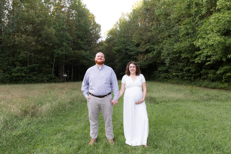 Expecting parents standing in a green field during their professional maternity photography session in Huntsville, Alabama. Mother is wearing a white dress, holding her belly expectantly.